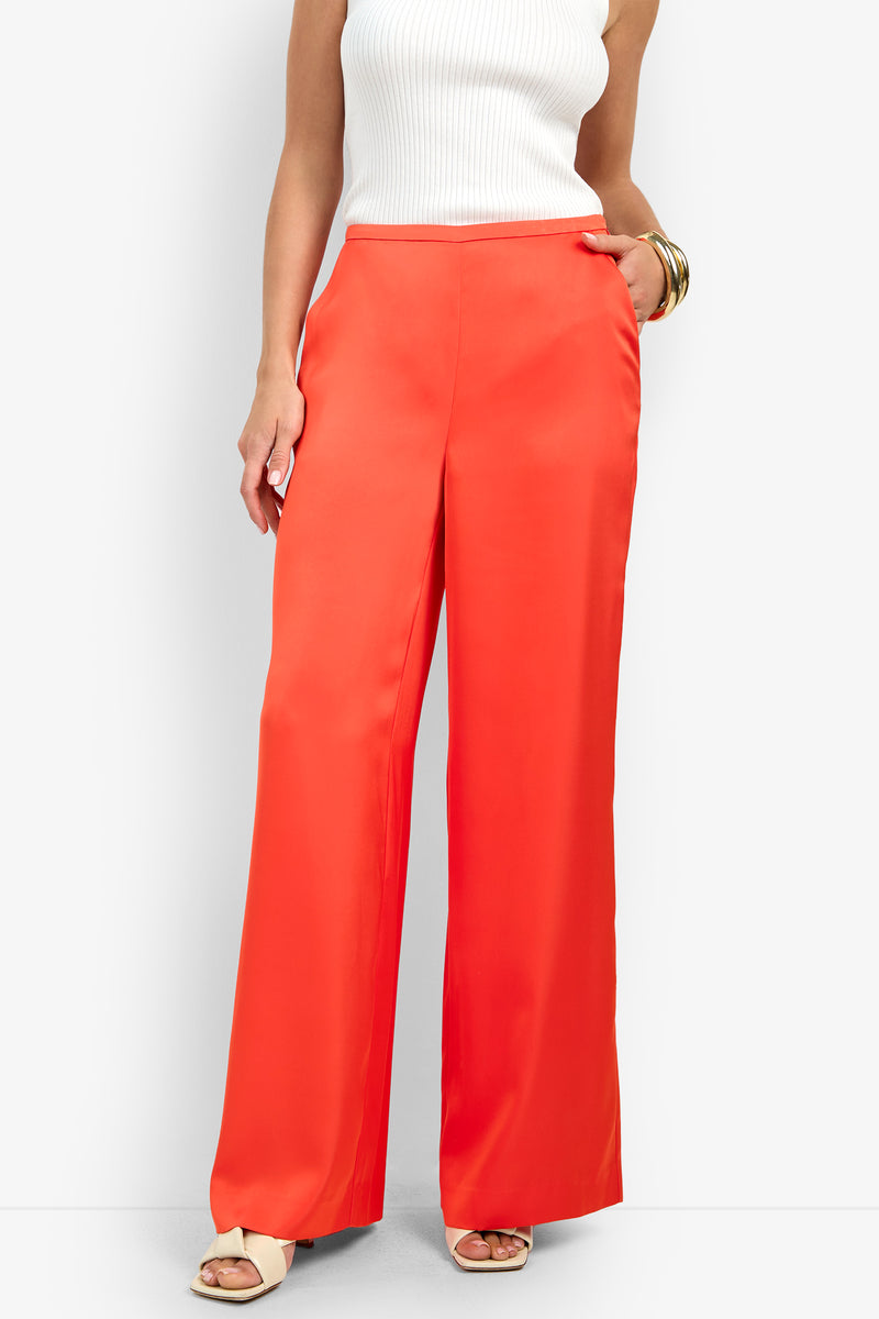 A woman wearing the TAVIA PANT in bright orange with a white sleeveless ribbed top, beige open-toe heels, and gold bangles stands with one hand in her pocket.
