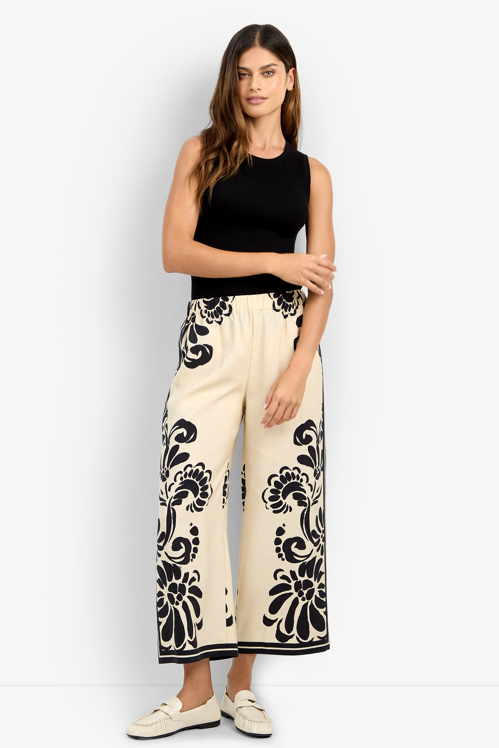 A woman with long brown hair stands against a plain white background, wearing a sleeveless black top, cream loafers, and the ESTELLA 2 PANT—cream wide-leg pants featuring bold black floral patterns.