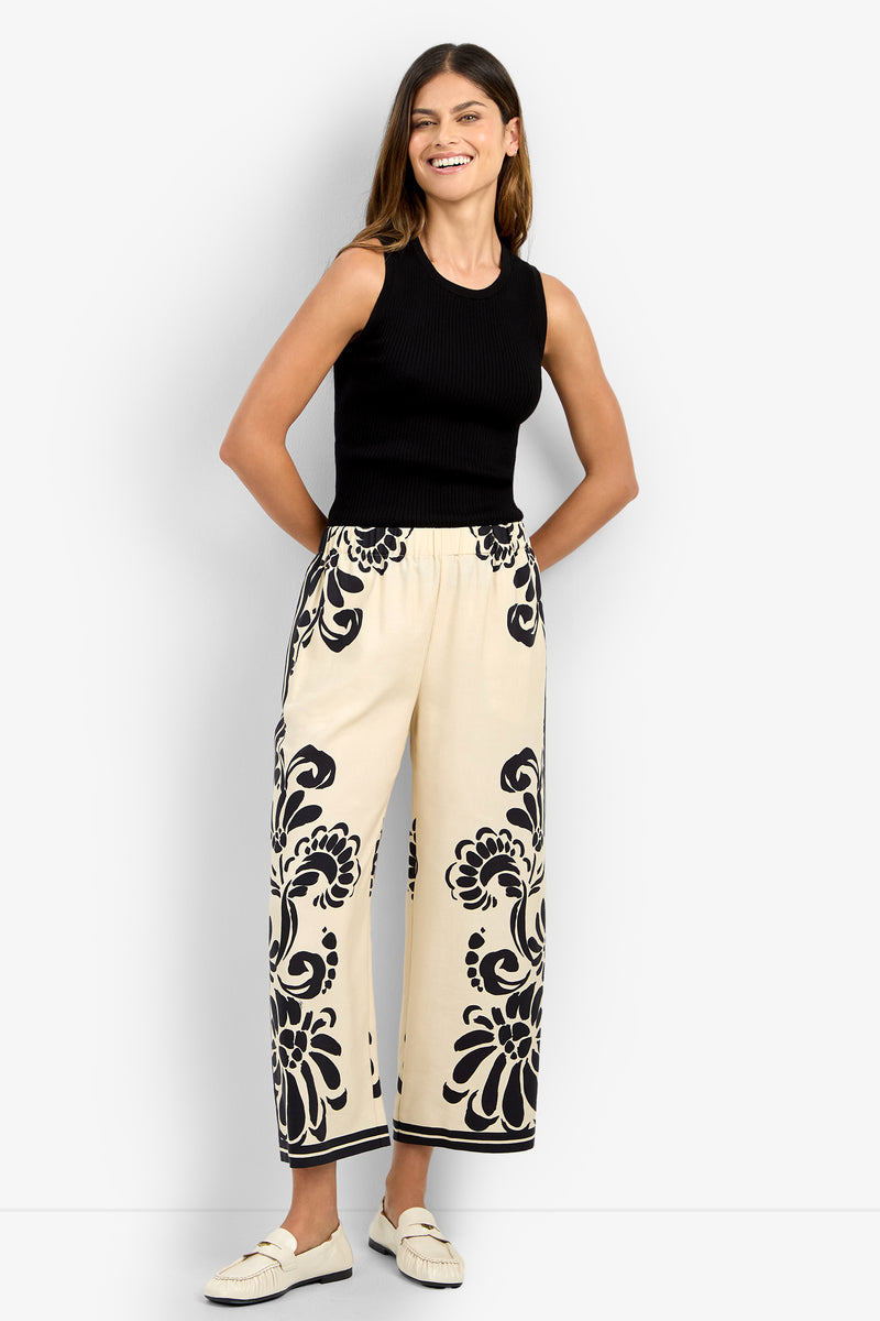 A woman smiles wearing the ESTELLA 2 PANT—cream wide-leg pants with bold black floral patterns on the sides and hem—paired with a sleeveless black top and cream loafers.
