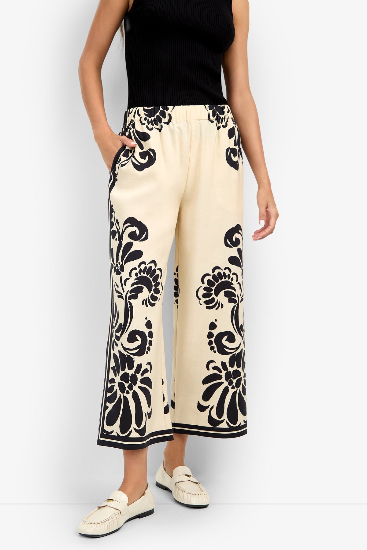 Wearing the ESTELLA 2 PANT in cream with bold black florals, a sleeveless black top, and cream loafers, this person stands confidently with one hand in their pocket.