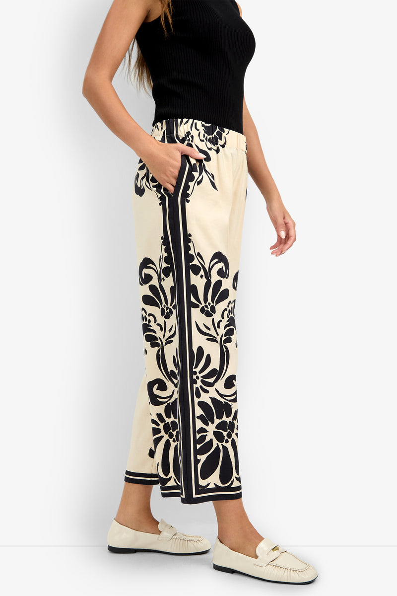 Woman wears the ESTELLA 2 PANT—cream-colored pants with bold black floral patterns and stripes—paired with a black sleeveless top and cream loafers, standing sideways with one hand in her pocket.