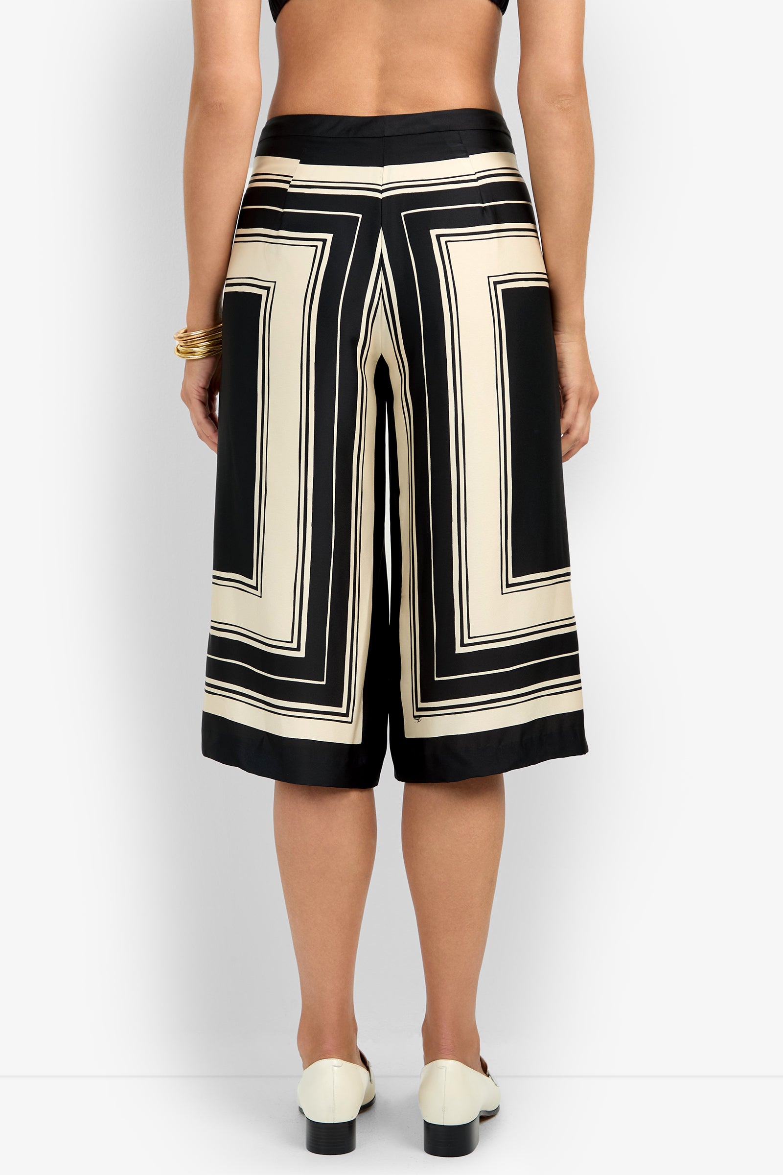 A person shown from the back against a plain white background wears black and cream geometric patterned LANAY CROP PANT culotte shorts, a black top, white shoes, and gold bangles.