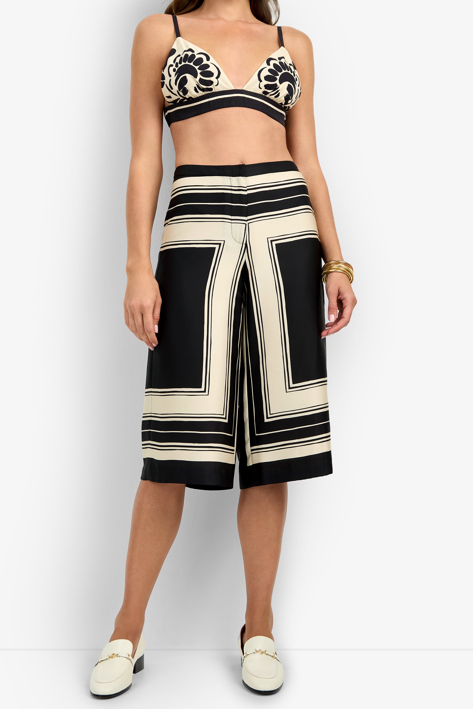 A woman wears the LANAY CROP PANT—cream and black patterned wide-leg culottes—with a matching bralette, white loafers, and gold bracelets, posed against a plain white background.