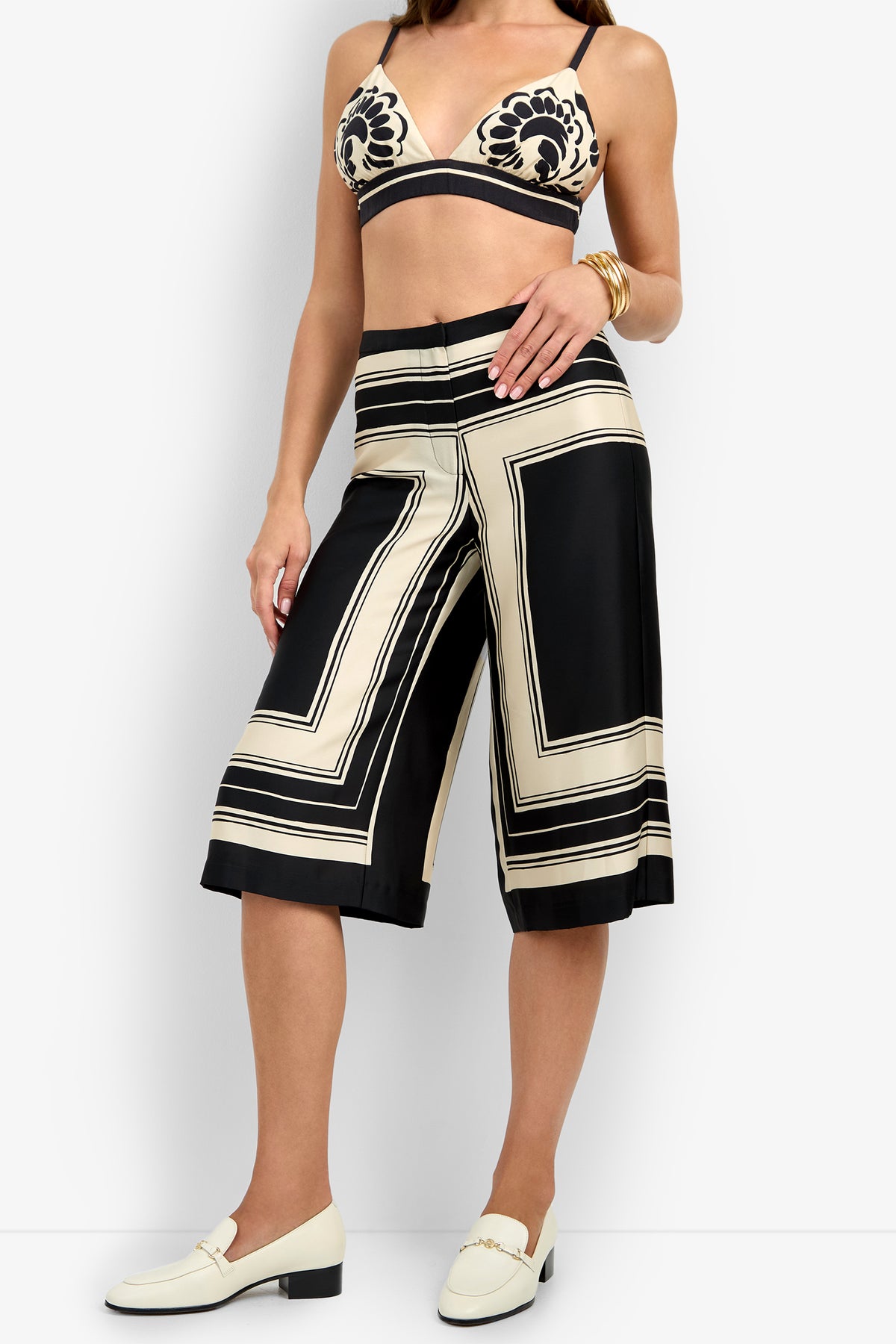 A woman wears a beige and black floral bralette, white loafers, gold bangles, and the LANAY CROP PANT—high-waisted wide-leg culottes with geometric square patterns.