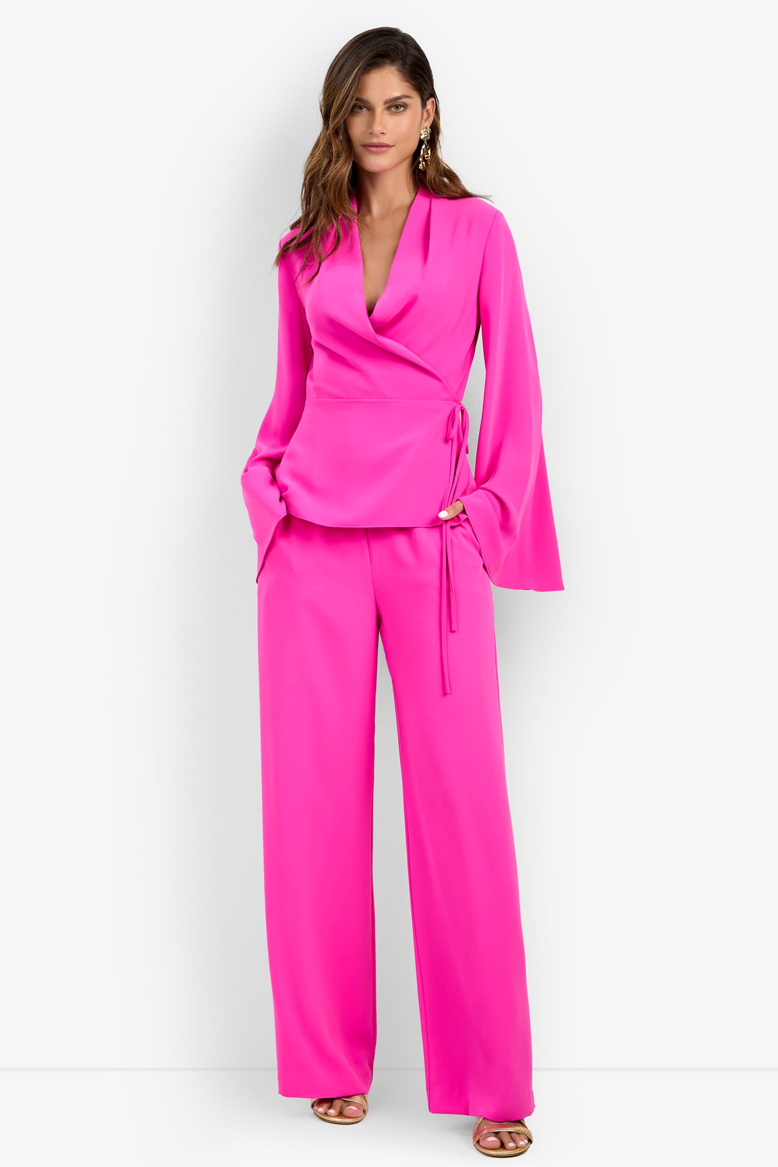 A woman stands with her hands in the pockets of the Tavia Pant in Luminosa Rosa, paired with a matching bright pink wrap blouse with wide sleeves, against a white background.