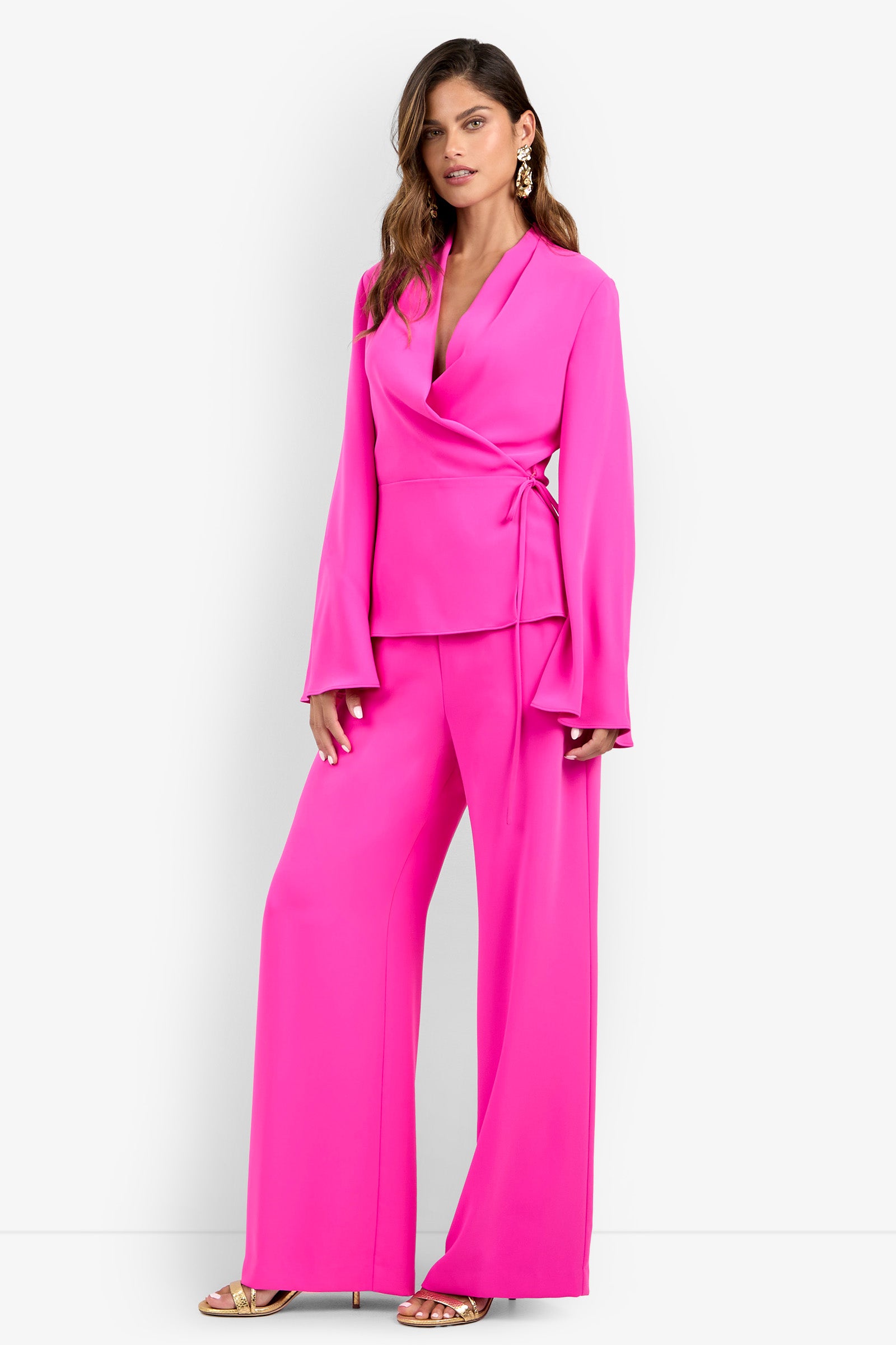 A woman stands against a plain white background wearing the bright pink Tavia Pant with a matching long-sleeve wrap blouse, gold sandals, and statement earrings.