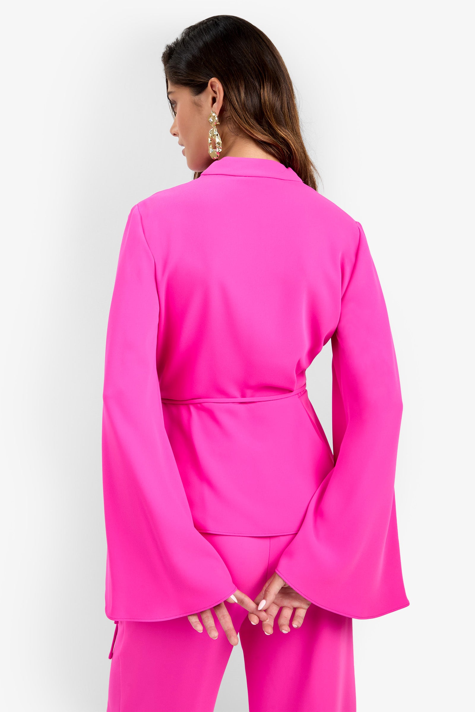 A woman stands with her back to the camera against a plain white background, wearing the Tavia Pant as part of a bright pink suit with wide sleeves, her hands clasped behind her back, and accessorized with gold statement earrings.
