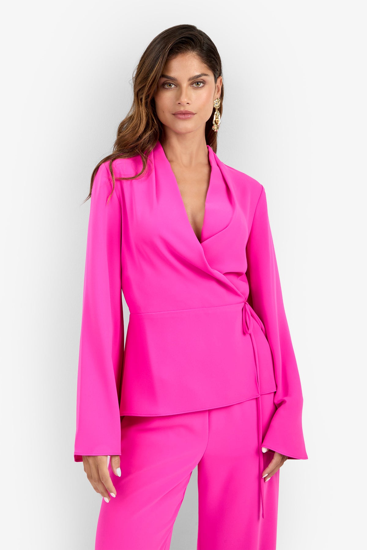 A woman stands against a plain white background wearing the Tavia Pant styled with a bright pink long-sleeve wrap blazer. Her long wavy hair is complemented by gold earrings.