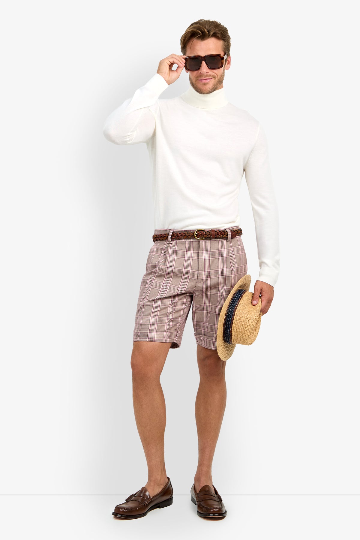 A man wearing sunglasses, a white turtleneck, OLIVER PLEATED SHORTS, a braided belt, and brown loafers stands against a plain white background holding a straw hat with a blue band in his left hand.