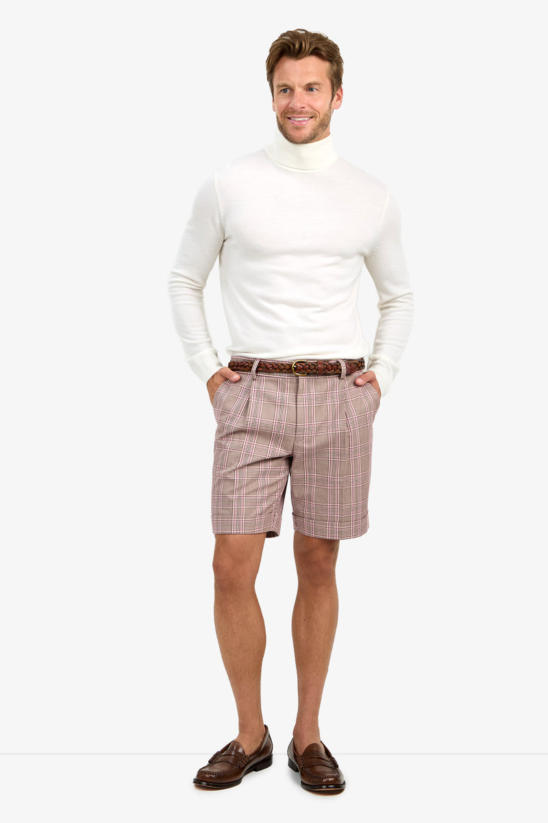 A man stands smiling against a light background, wearing a white turtleneck sweater, brown loafers, and the OLIVER PLEATED SHORT in pink and white plaid with a brown belt.