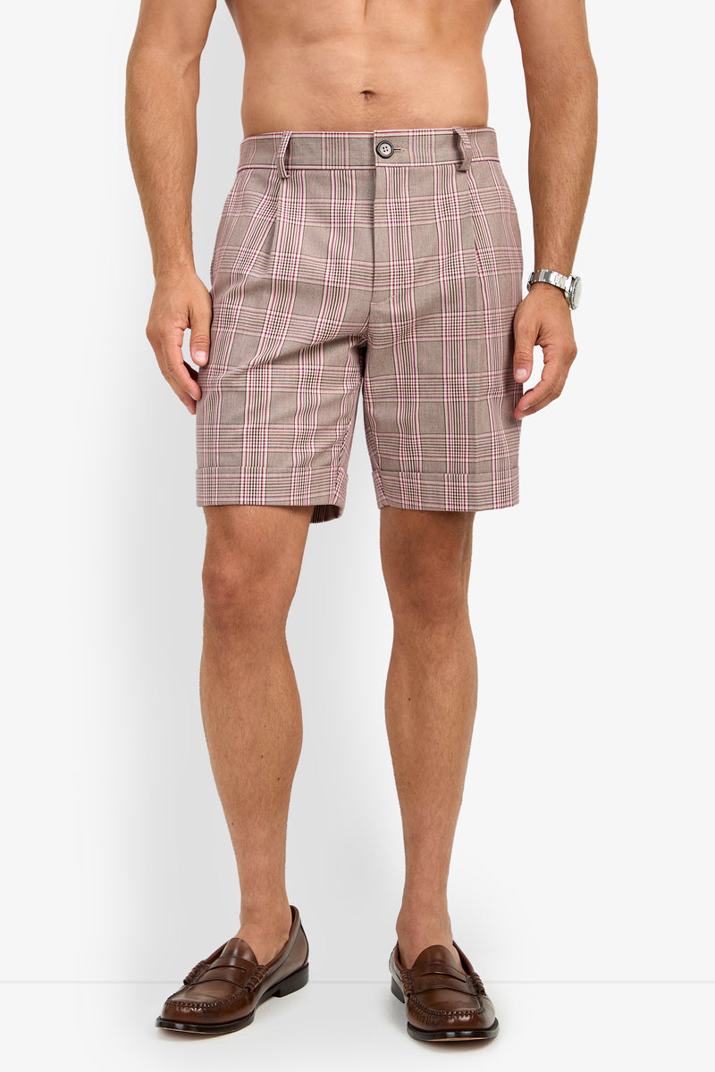 A man stands against a plain white background wearing the OLIVER PLEATED SHORT in brown plaid with brown loafers and a watch. His upper body is bare, and his hands are relaxed at his sides.