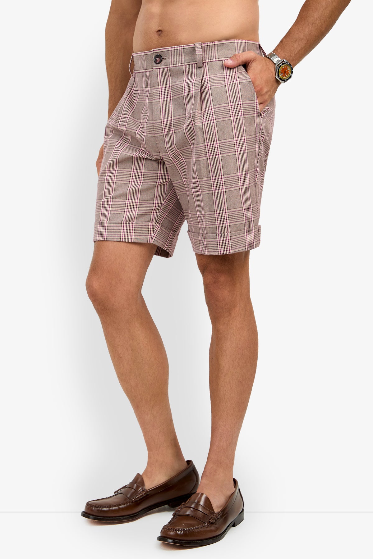 A man wears the OLIVER PLEATED SHORT, paired with brown loafers and a watch, standing with one hand in his pocket against a white background.