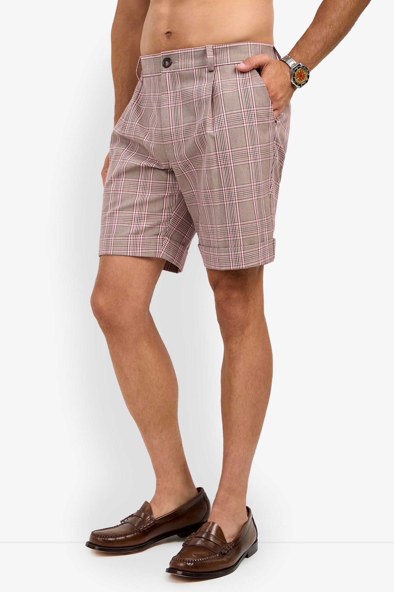 A man wears the OLIVER PLEATED SHORT, paired with brown loafers and a watch, standing with one hand in his pocket against a white background.