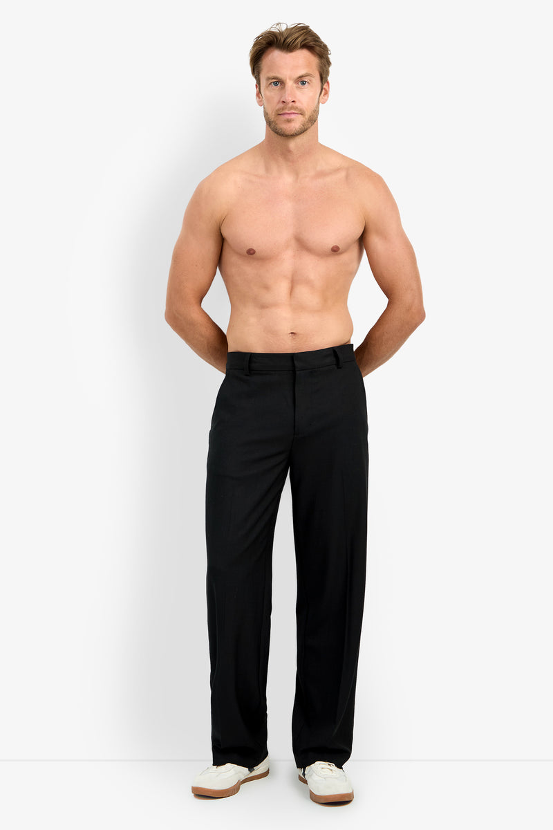 A shirtless man with light skin and short hair stands facing forward against a plain white background, wearing the BUCKINGHAM TROUSER in black with white sneakers. His hands are behind his back.