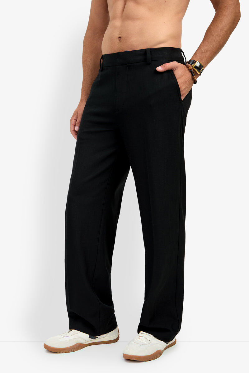 A shirtless model showcases the BUCKINGHAM TROUSER—a pair of black dress pants—paired with white sneakers, standing against a plain background with one hand in their pocket.