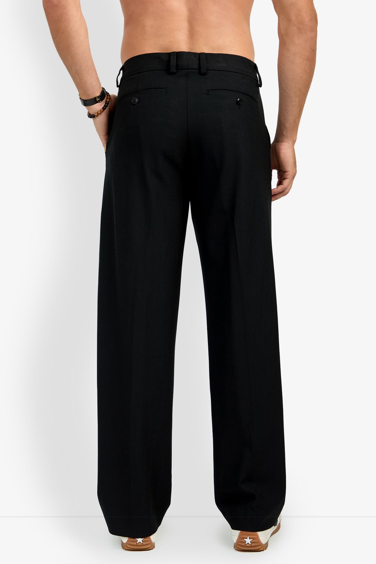 A shirtless person stands facing away, wearing loose-fitting BUCKINGHAM TROUSER in black and brown sneakers with white accents, against a plain white background.