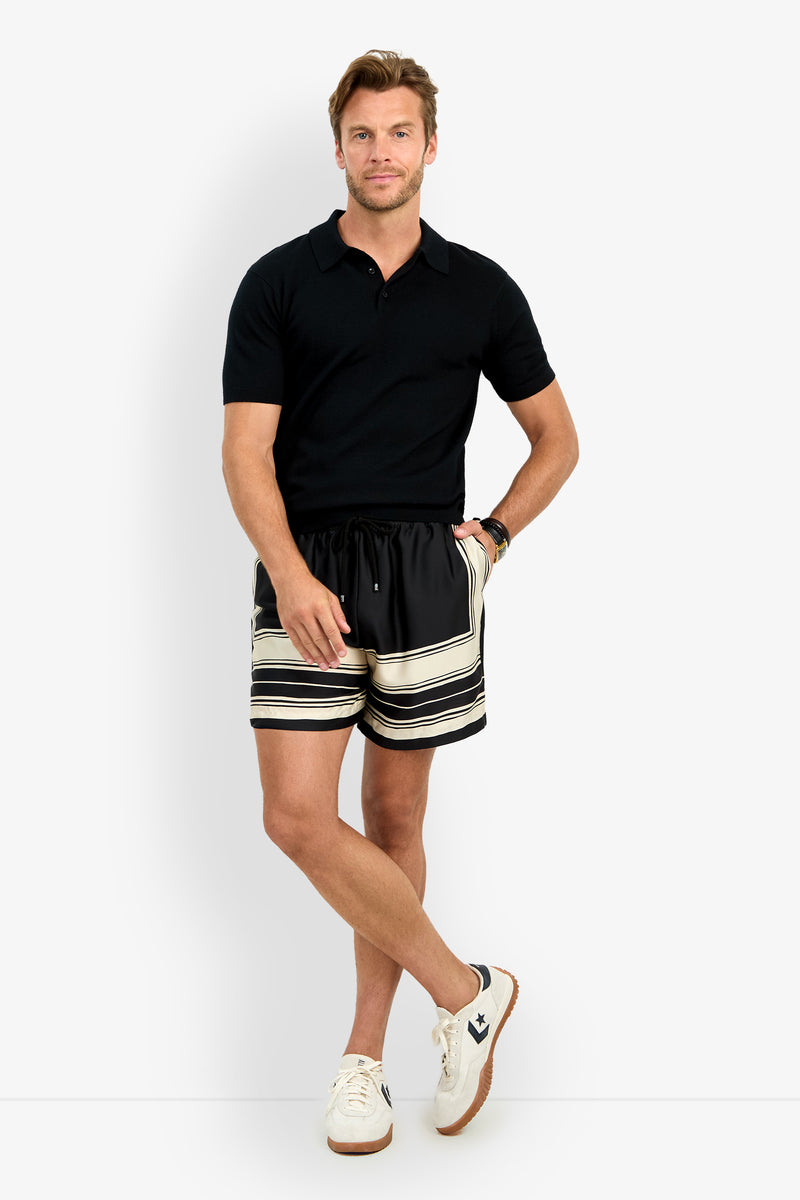 A man with short, light brown hair stands against a white background, one leg crossed over the other, wearing a black polo, SHORELINE SHORT in black and beige stripes, white sneakers, and a watch.