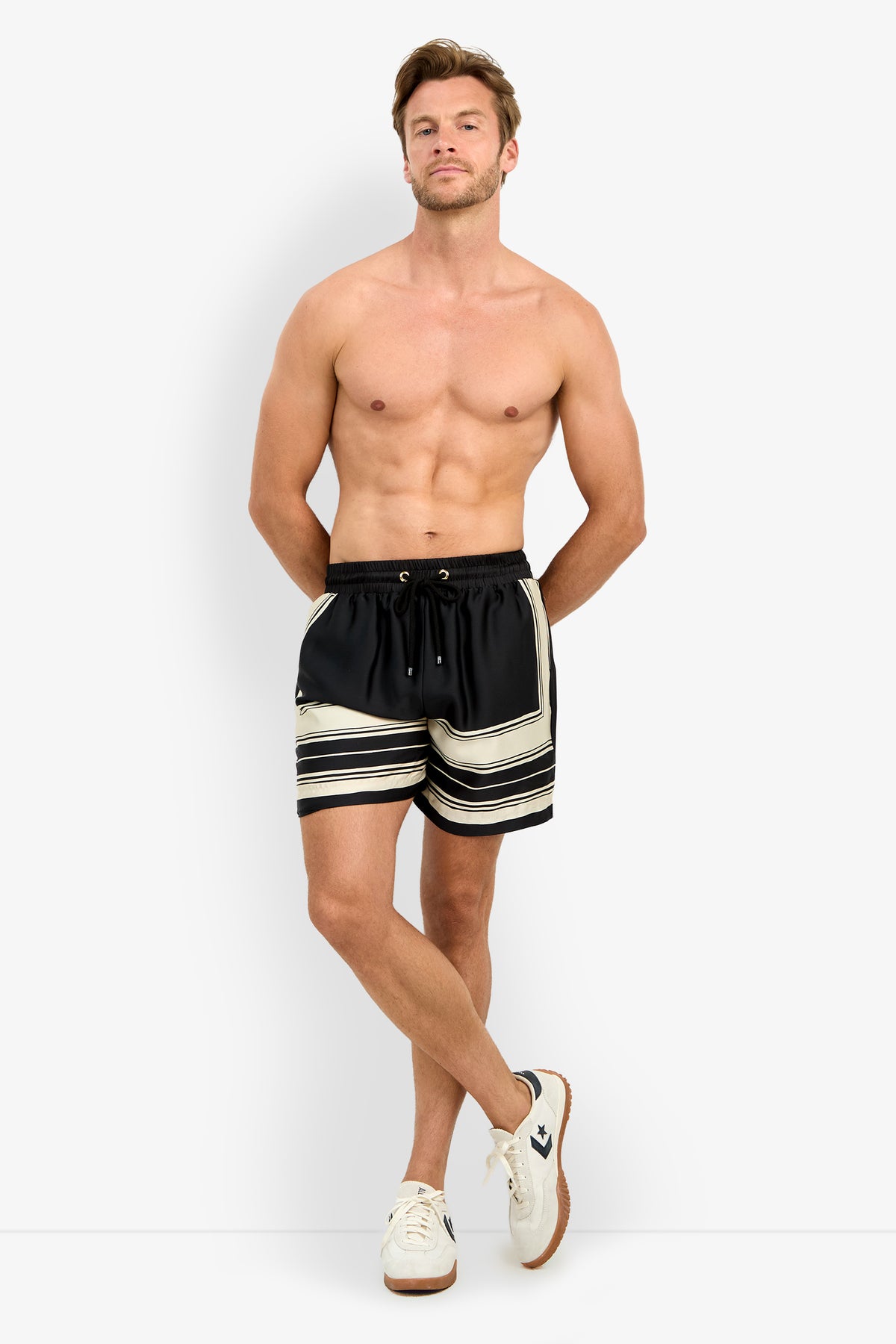 A shirtless man stands against a plain white background, wearing the SHORELINE SHORT in black and beige stripes with white sneakers. His hands are behind his back as he looks confidently at the camera.