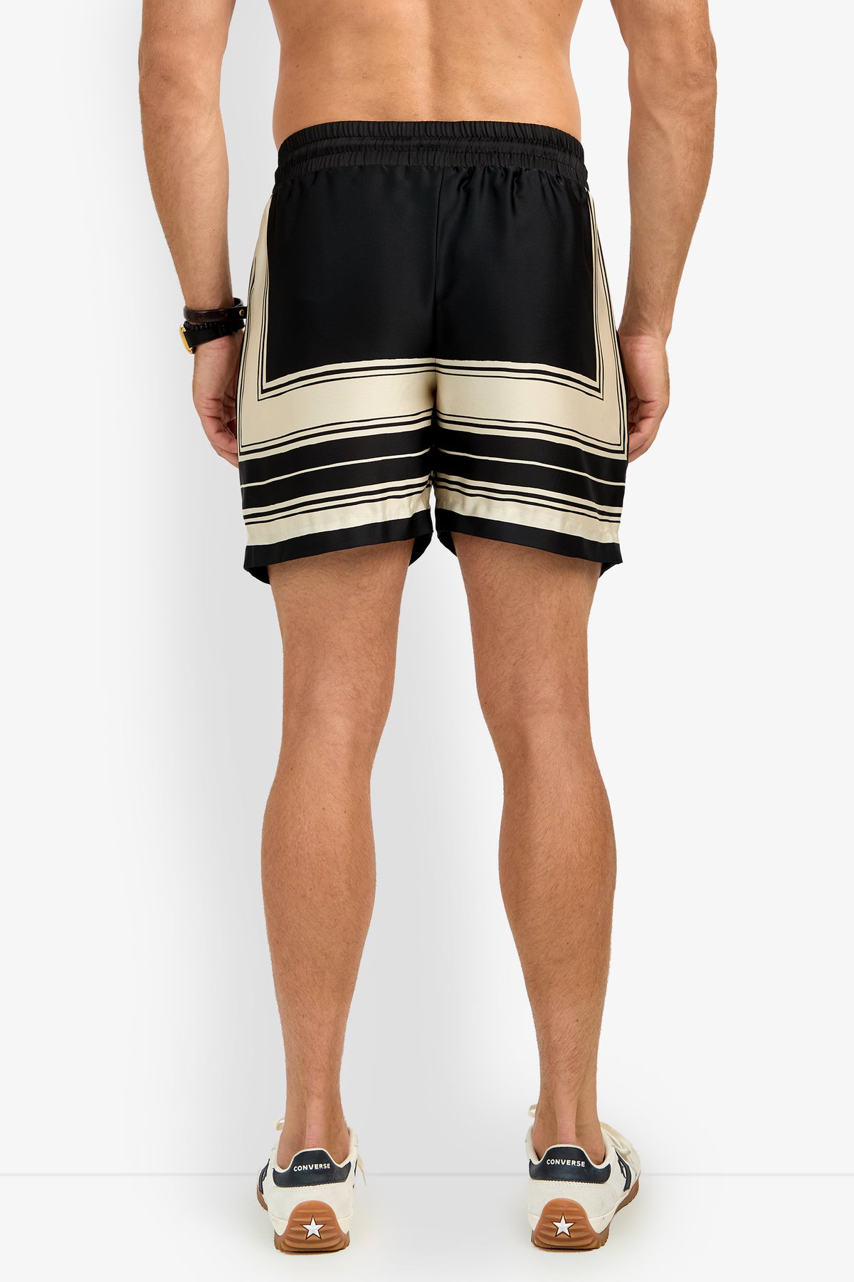 A man stands shirtless against a plain white background, wearing SHORELINE SHORT black and beige patterned shorts, a beaded bracelet, and white sneakers with brown soles.