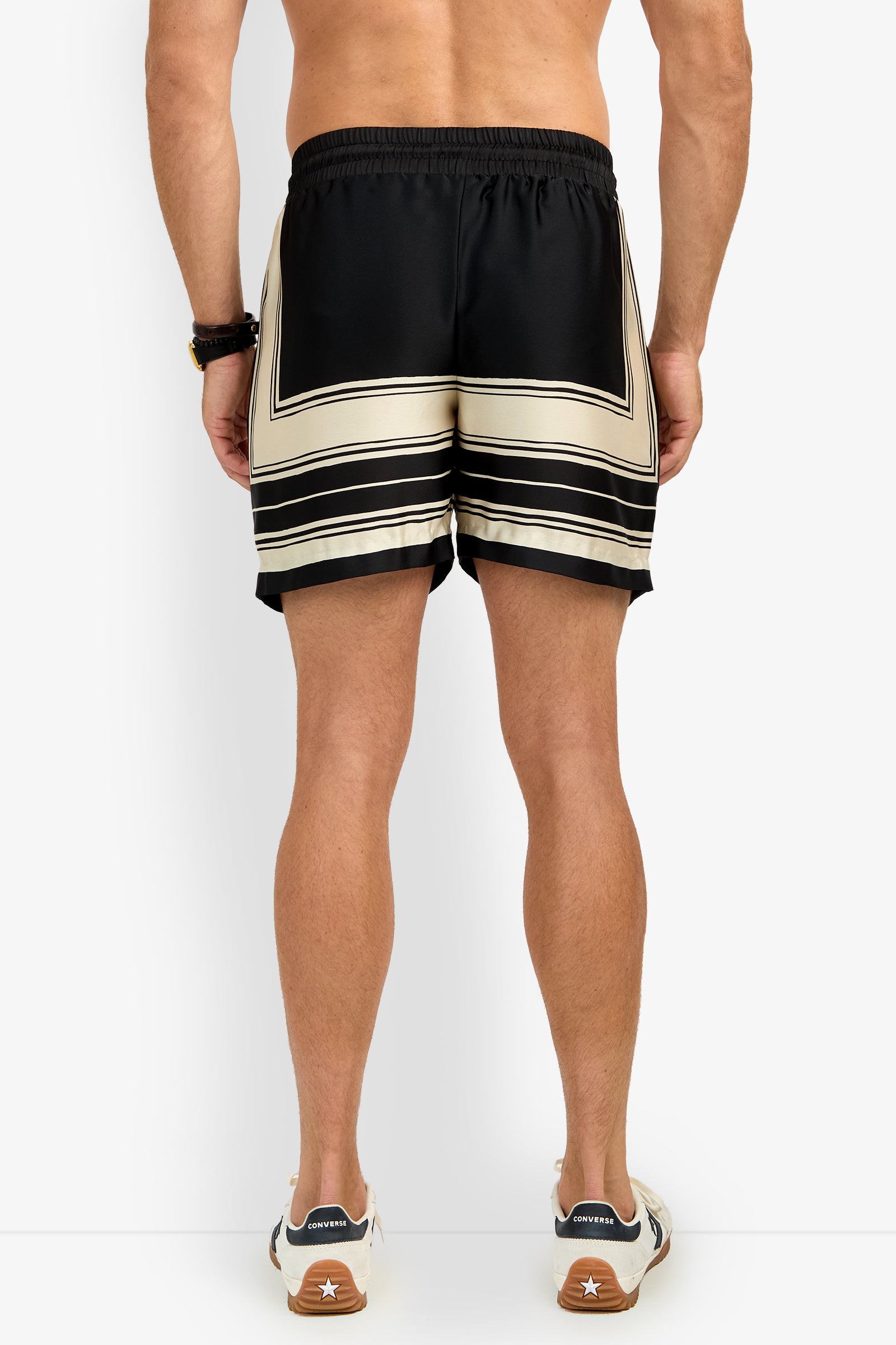 A man stands shirtless against a plain white background, wearing SHORELINE SHORT black and beige patterned shorts, a beaded bracelet, and white sneakers with brown soles.