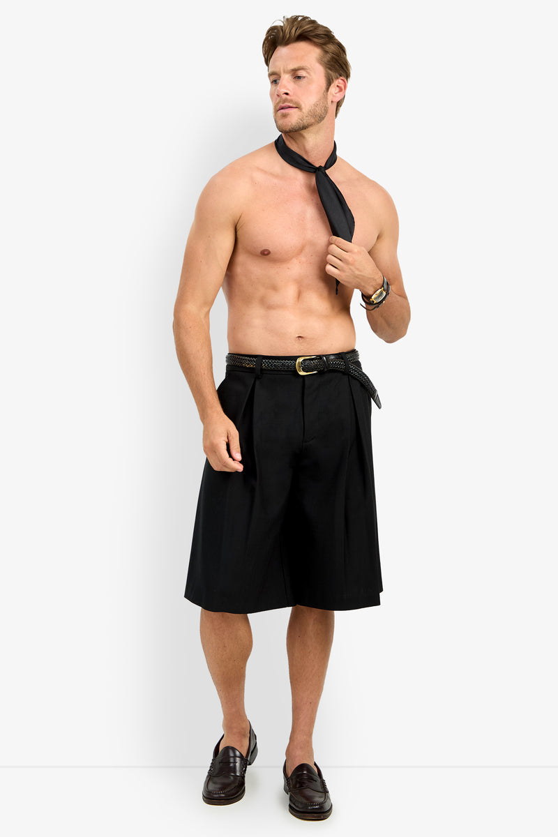 A shirtless man with a loosened black tie stands against a plain background, wearing the GALFANO WIDE LEG SHORT, a belt, a watch, and dark loafers. He gazes to the side with a relaxed expression.