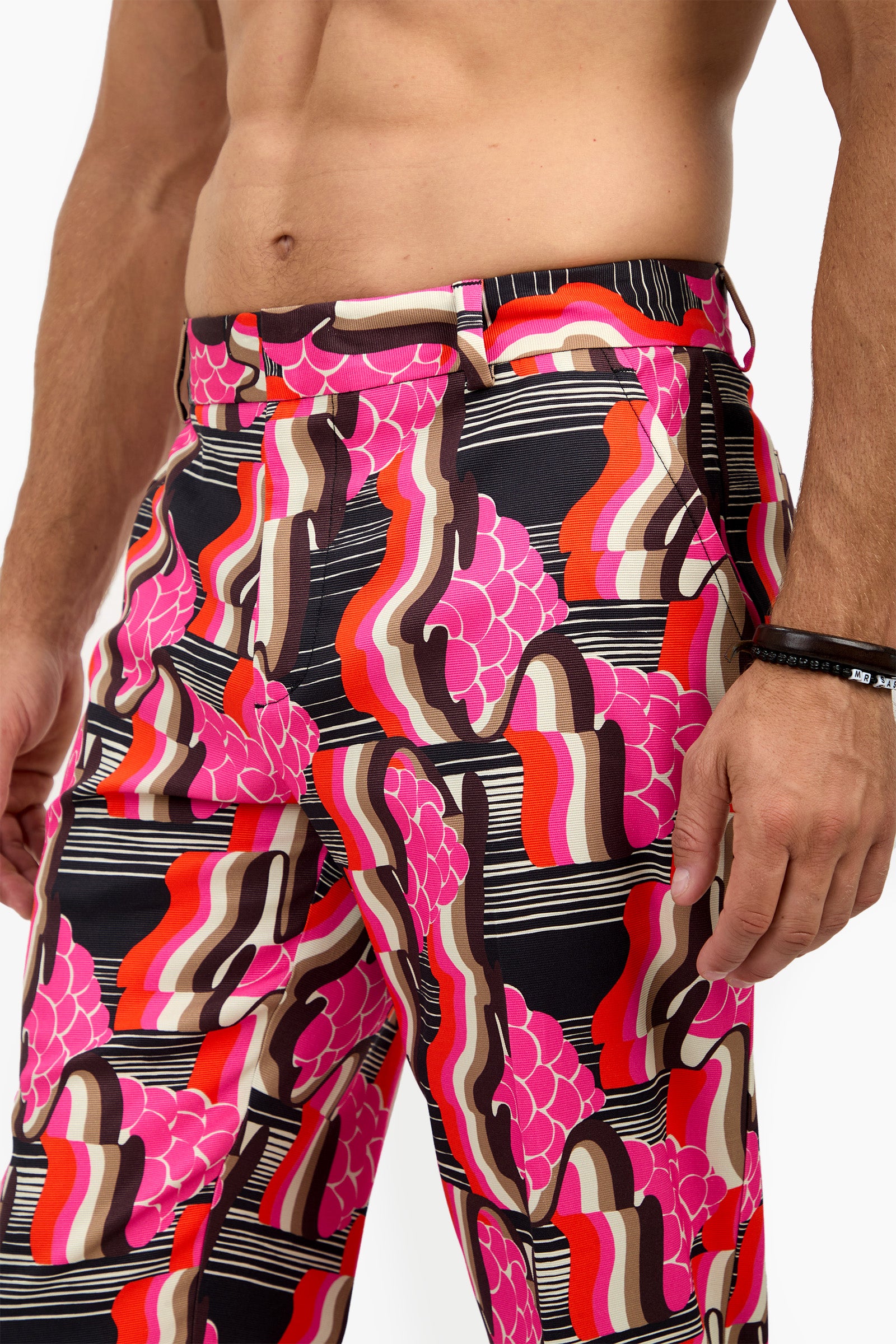 A person wearing the CLYDE SLIM TROUSER—high-waisted pants with a bold, retro geometric pattern in pink, red, brown, black, and white—stands against a white background with a bare torso.