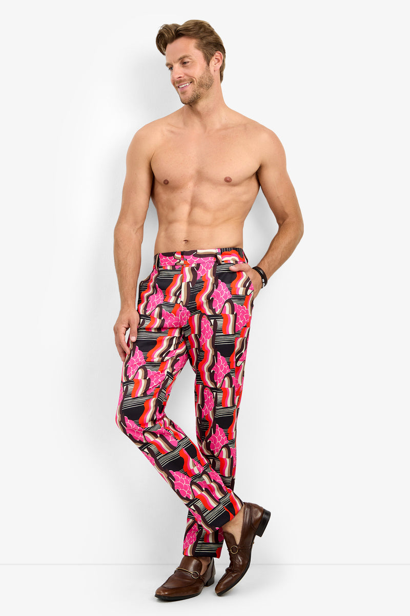A shirtless man stands against a white background, wearing the CLYDE SLIM TROUSER featuring a vibrant pink, red, and black lipstick print pattern and brown dress shoes. He smiles with his arms relaxed at his sides.