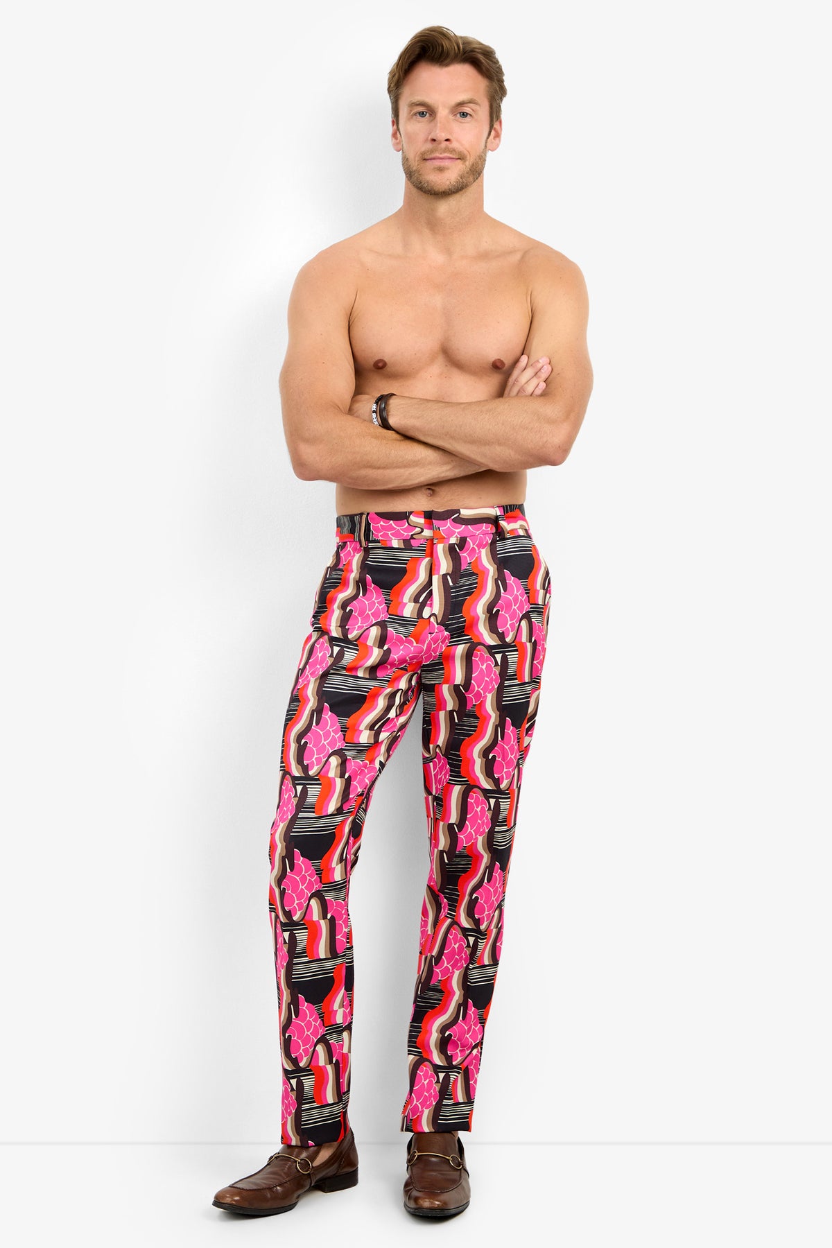 A man stands barefoot against a plain background, shirtless with arms crossed, wearing the CLYDE SLIM TROUSER featuring bold pink, red, and black abstract patterns.