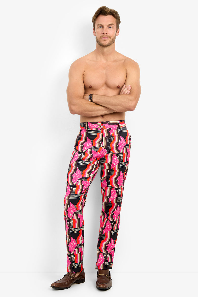 A man stands barefoot against a plain background, shirtless with arms crossed, wearing the CLYDE SLIM TROUSER featuring bold pink, red, and black abstract patterns.