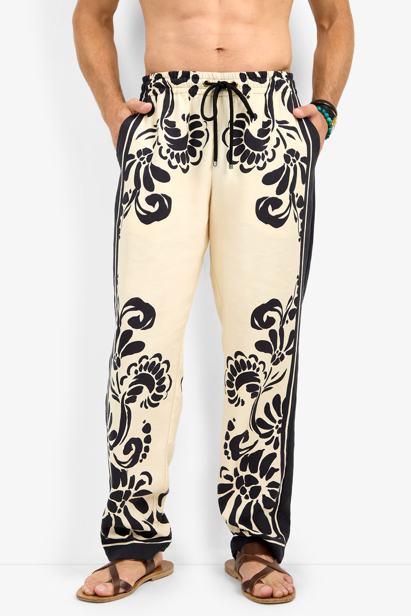 A man wears the FELIPE DRAWSTRING PANT, featuring cream fabric with bold black floral patterns and black side stripes. He pairs them with brown sandals, has his hands in his pockets, and is shirtless.