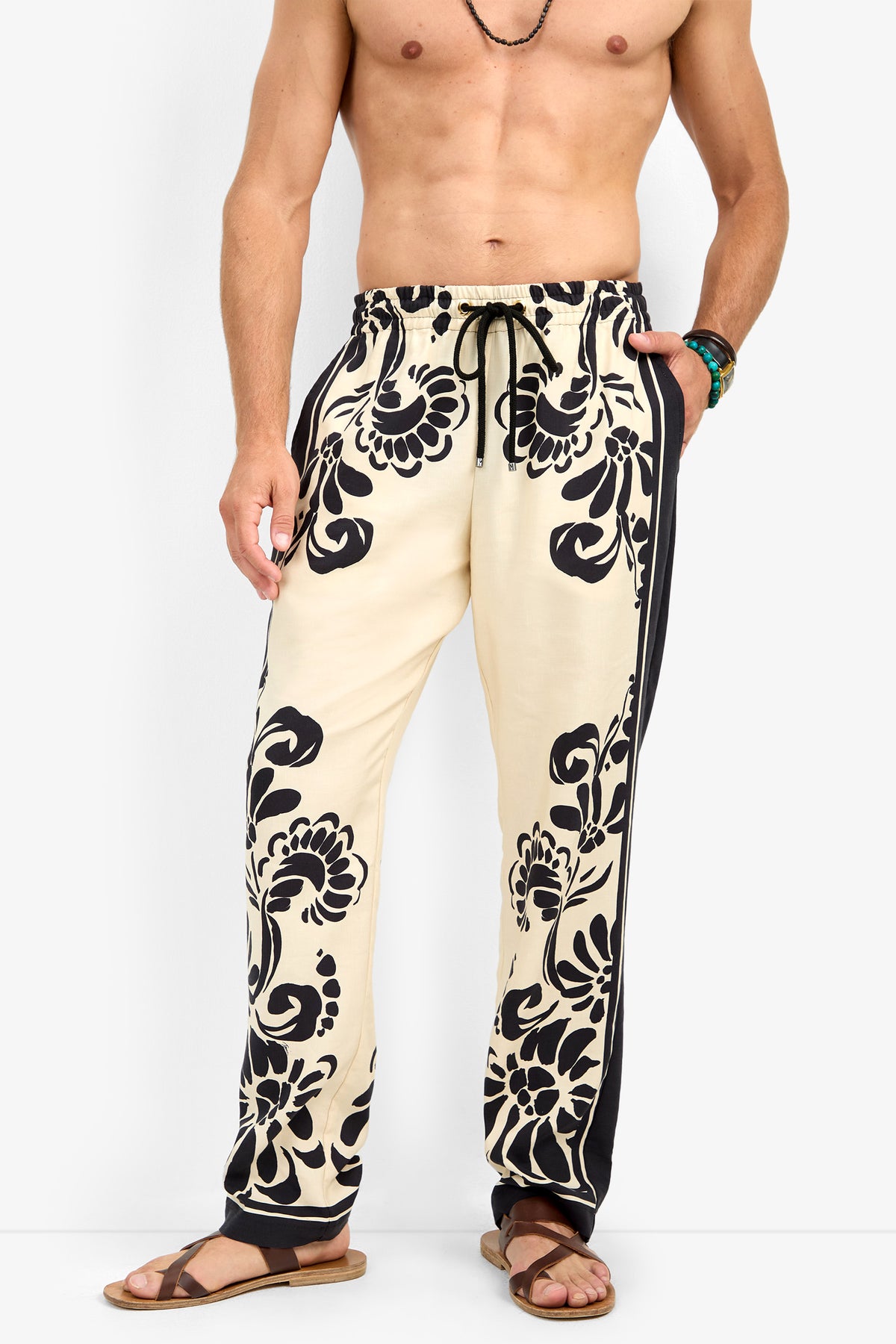 A shirtless man wearing the FELIPE DRAWSTRING PANT—cream-colored with bold black floral patterns and black side panels—paired with brown sandals, a necklace, and a bracelet, stands against a plain white background.