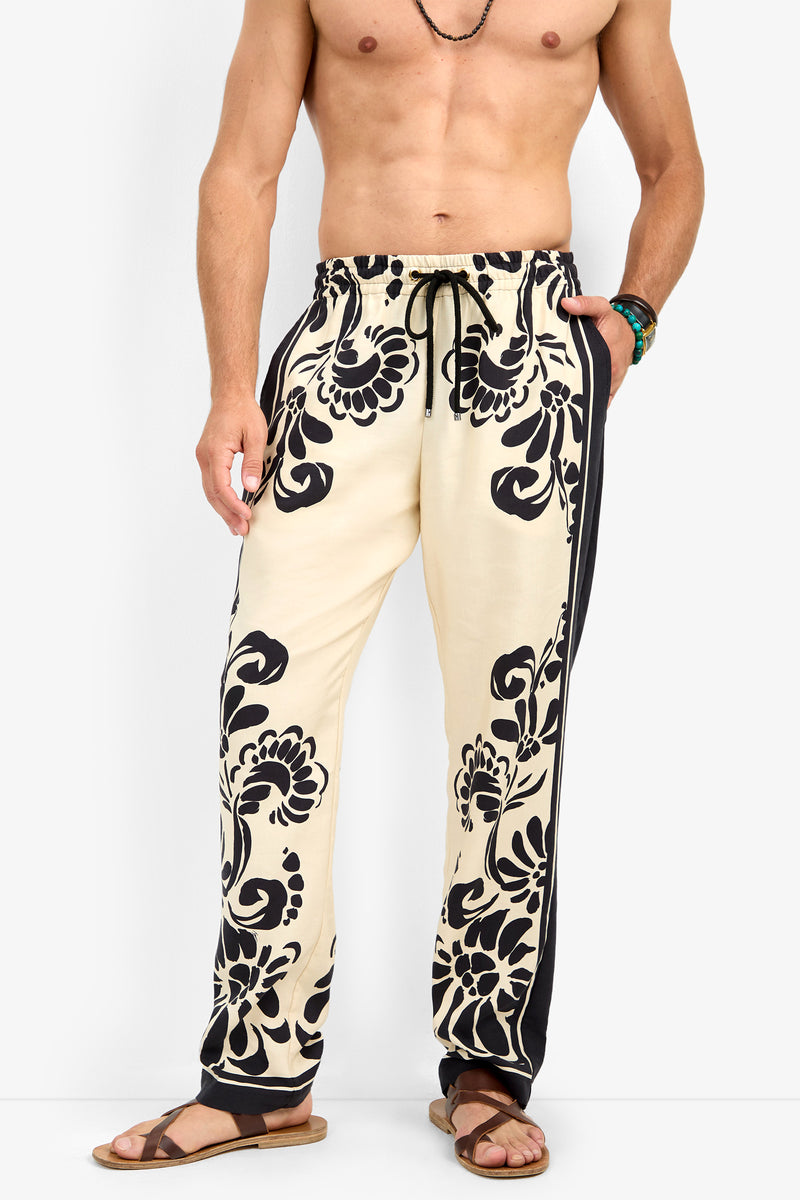 A shirtless man wearing the FELIPE DRAWSTRING PANT—cream-colored with bold black floral patterns and black side panels—paired with brown sandals, a necklace, and a bracelet, stands against a plain white background.