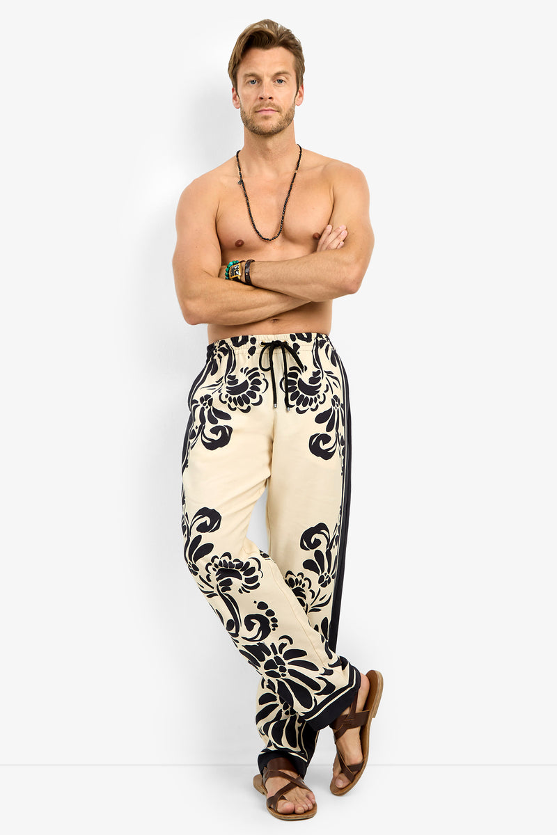 A man with light brown hair stands barefoot against a white background, arms crossed. He wears the FELIPE DRAWSTRING PANT with bold black floral patterns, a beaded necklace, bracelets, and brown sandals. His chest is bare.