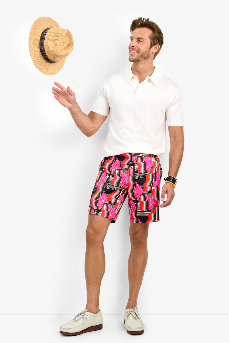 A man smiles against a white background, wearing colorful LAWRENCE SHORTS, a white polo shirt, and white shoes while tossing a straw hat in the air.