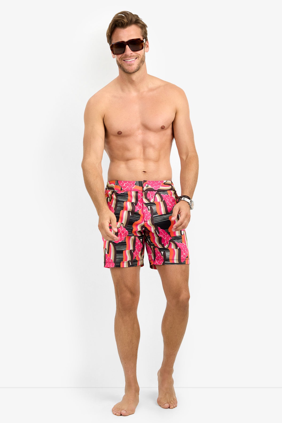 A smiling, barefoot man in sunglasses wears HONOLULU BOARDSHORT swim trunks with a pink lipstick tube pattern, standing against a plain white background.
