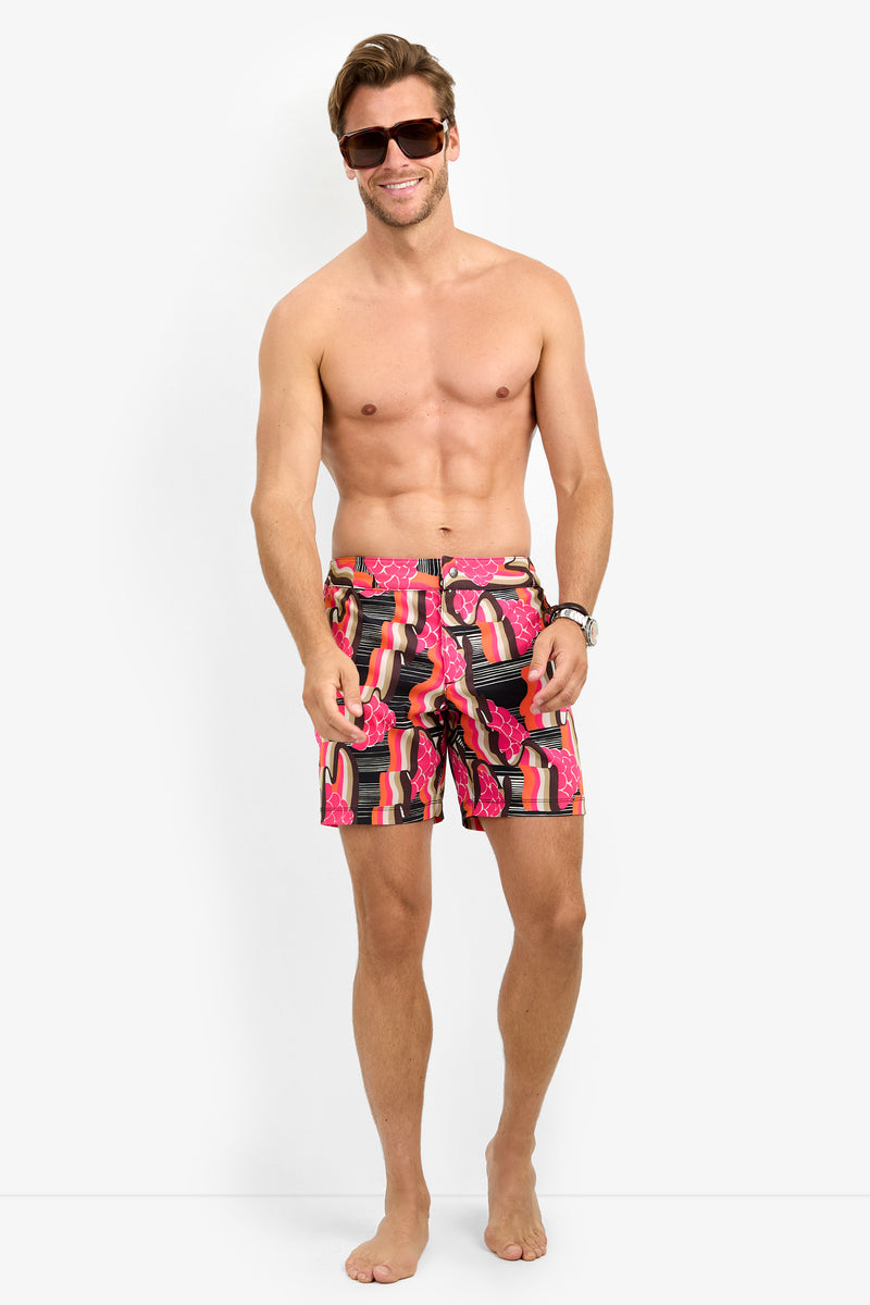 A smiling, barefoot man in sunglasses wears HONOLULU BOARDSHORT swim trunks with a pink lipstick tube pattern, standing against a plain white background.