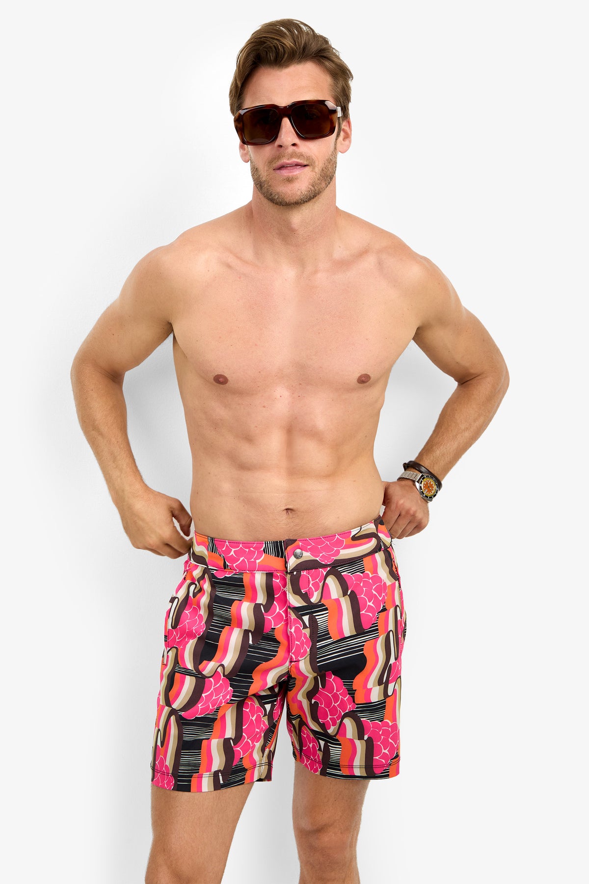 A shirtless man wearing large sunglasses and the HONOLULU BOARDSHORT in red, pink, black, and beige stands against a plain white background.
