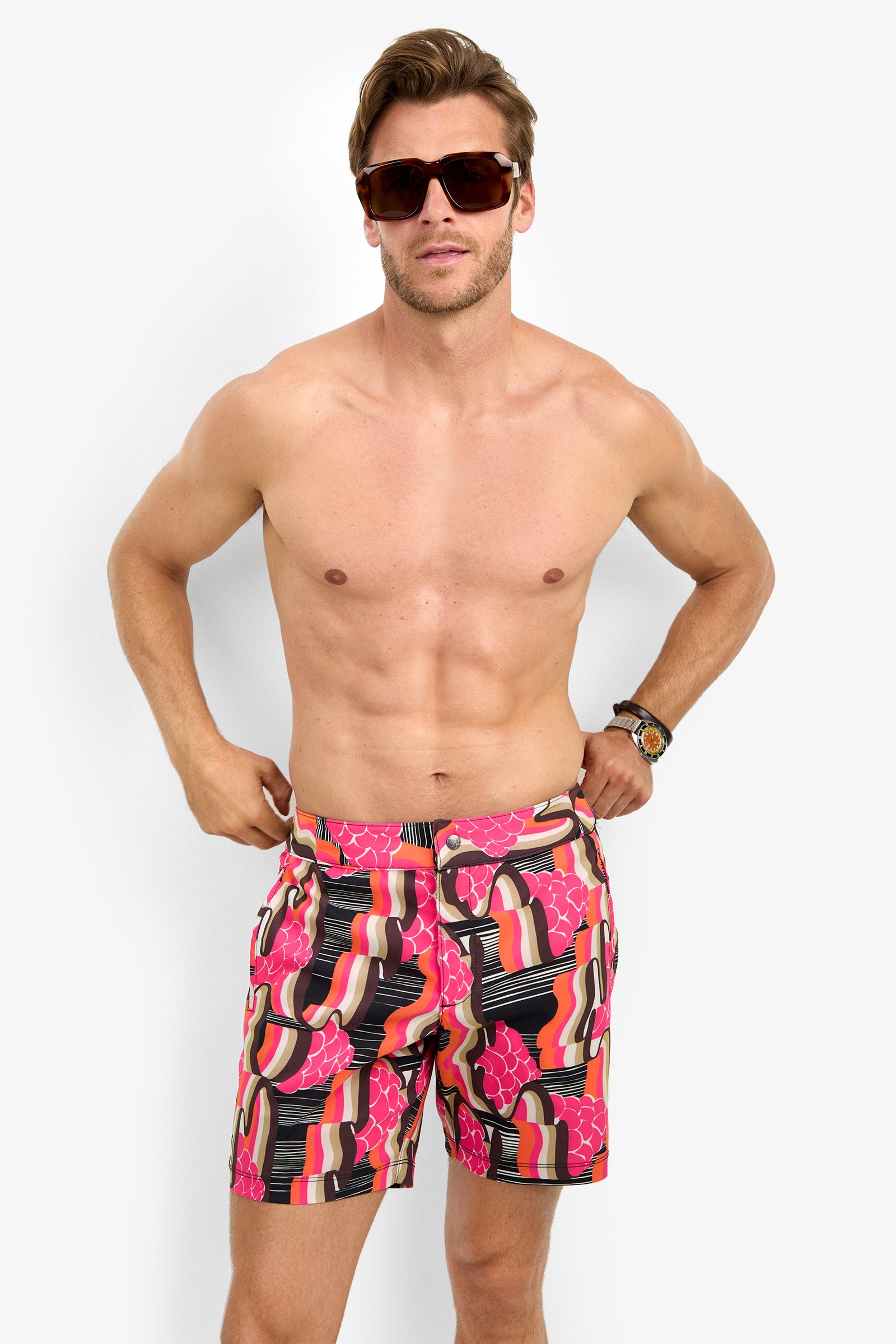 A shirtless man wearing large sunglasses and the HONOLULU BOARDSHORT in red, pink, black, and beige stands against a plain white background.