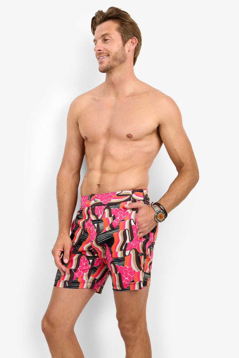 A smiling, shirtless man with light brown hair poses against a white background wearing the HONOLULU BOARDSHORT, which features a colorful pink and orange abstract pattern. He has one hand in his pocket and a watch on his left wrist.