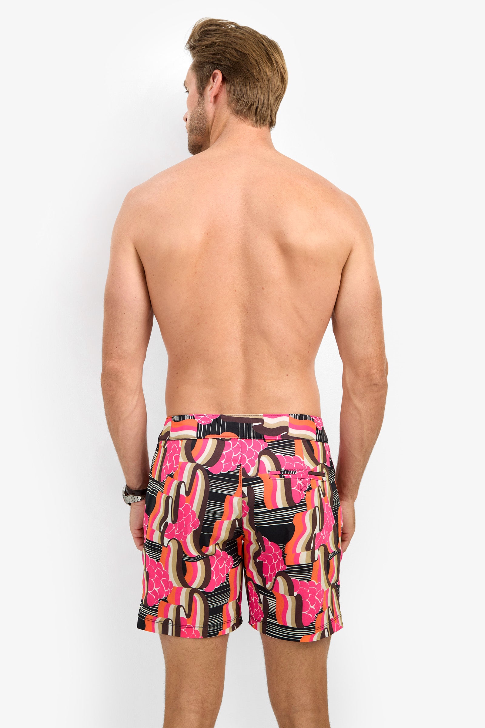 A shirtless man faces away, wearing the HONOLULU BOARDSHORT featuring pink, black, orange, and beige abstract patterns, set against a plain white background.