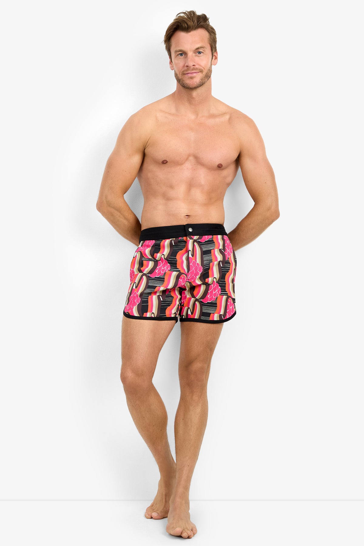 A smiling, shirtless man stands barefoot against a white background, wearing the SURFSIDE BOARDSHORT featuring a colorful red, pink, and black pattern. He has one leg crossed in front of the other.
