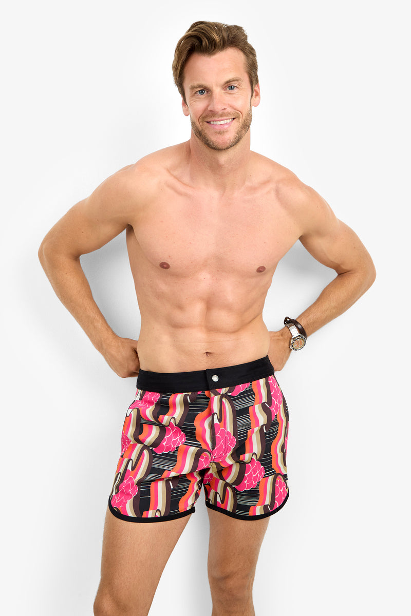 A smiling man stands against a white background, wearing the SURFSIDE BOARDSHORT with vibrant pink, red, and black patterns. He is shirtless, posing confidently with one hand on his hip and the other by his side.