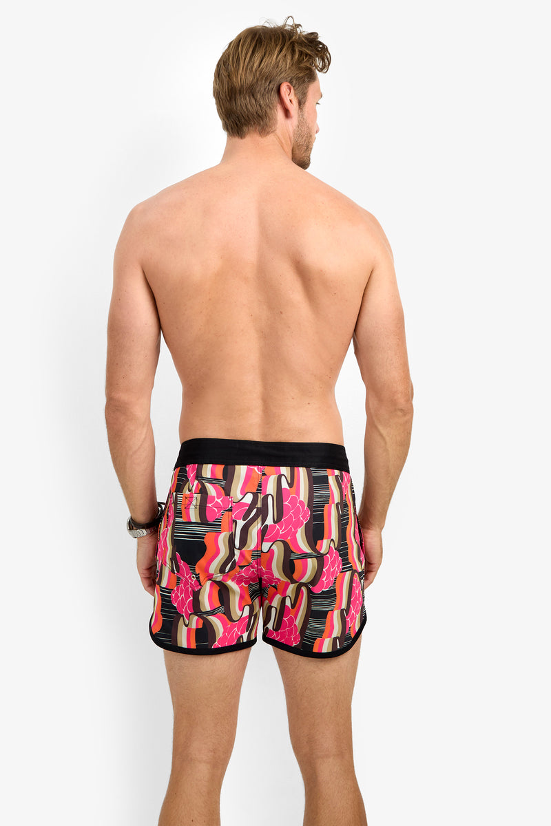 A light-skinned, brown-haired man stands shirtless with his back to the camera, wearing SURFSIDE BOARDSHORTS featuring pink, red, orange, and black patterns.