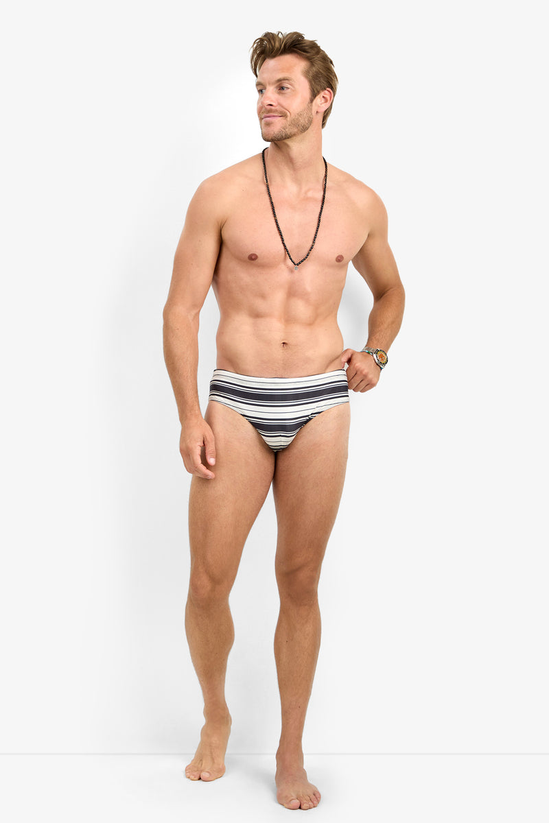 A shirtless man stands barefoot, wearing the BONDI SWIM BRIEF striped swimsuit and a black beaded necklace, looking to the side against a plain white background.