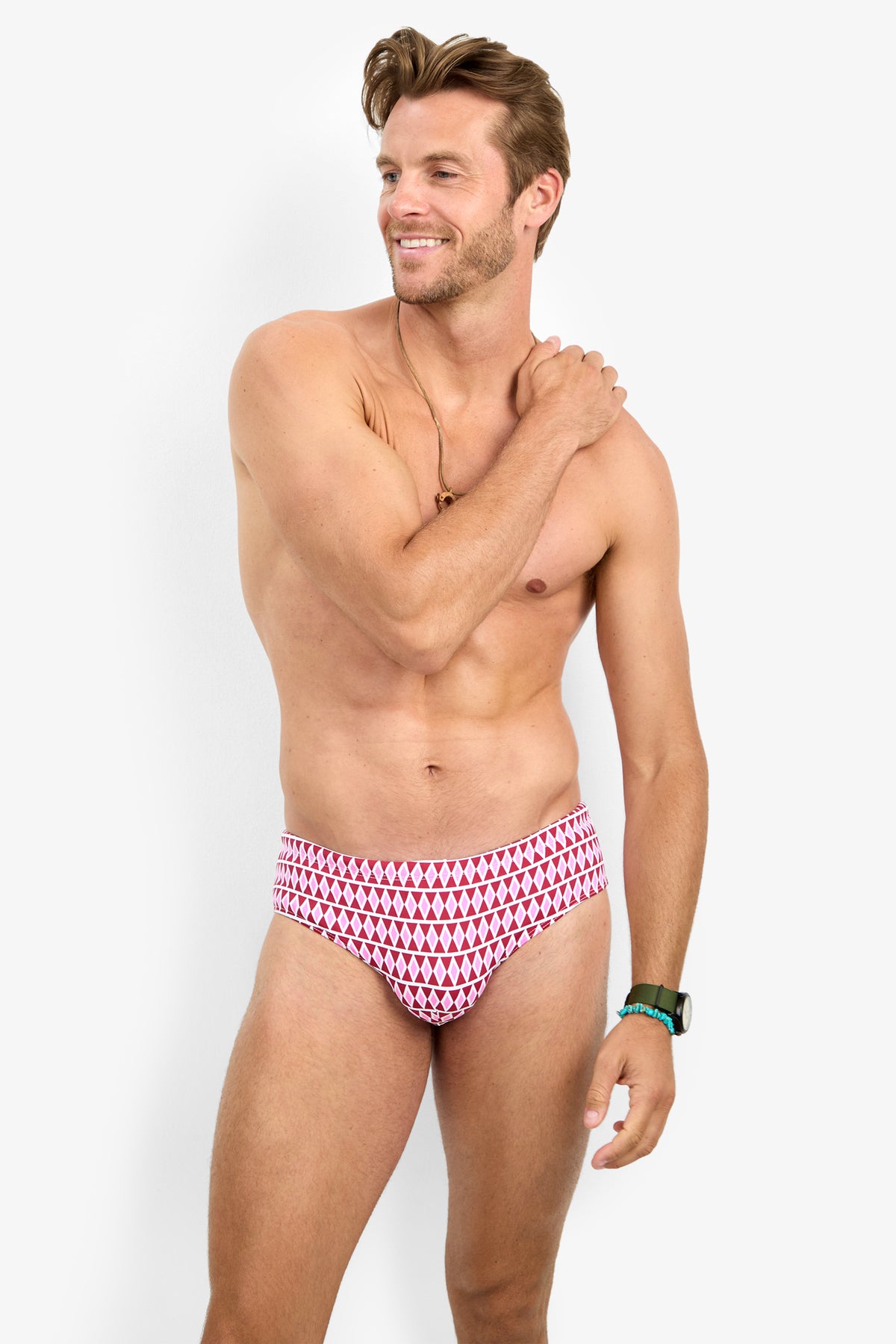 A smiling man with light brown hair stands against a white background, wearing the BONDI SWIM BRIEF, a watch, and a necklace, with one hand resting on his opposite shoulder.