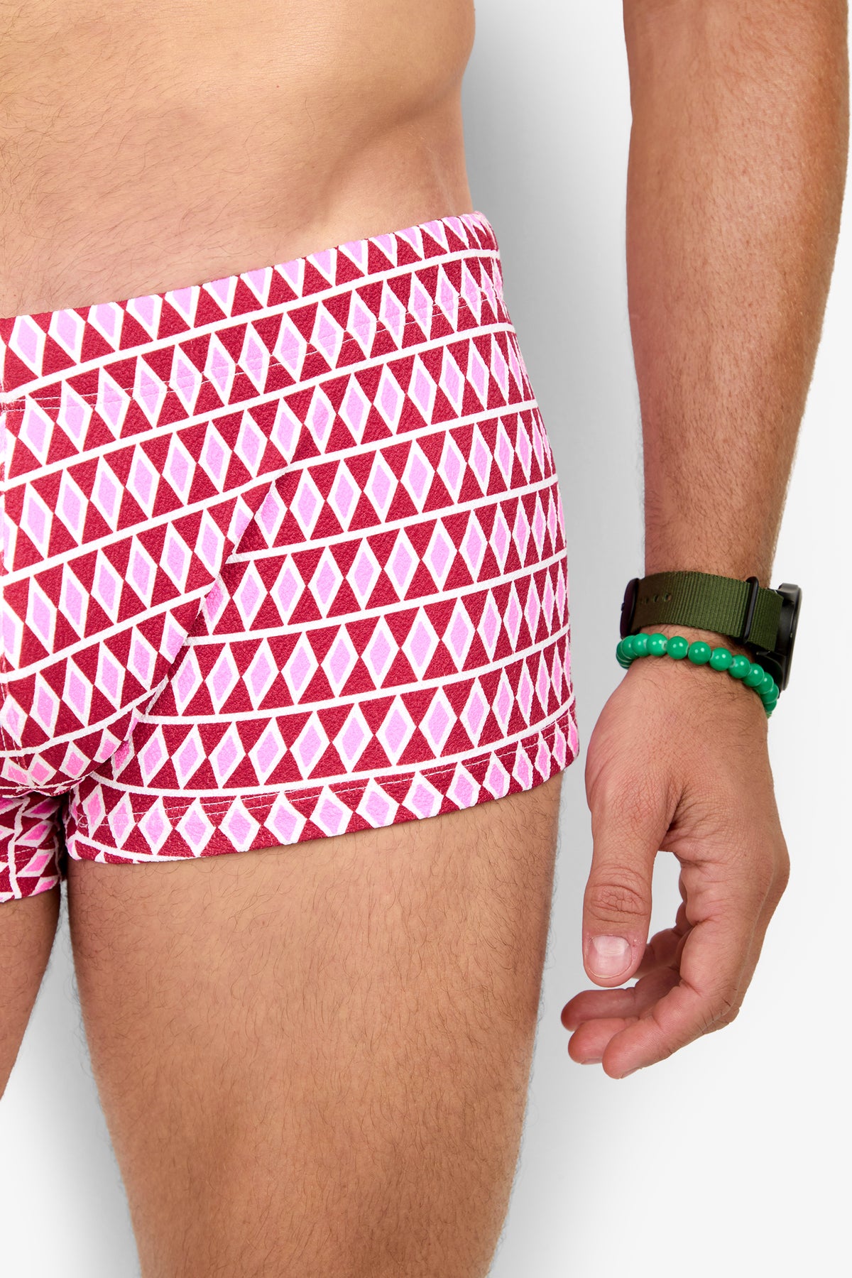 A person wears MONTEGO BAY SWIM TRUNK with a red and white geometric pattern, paired with a green watch and green beaded bracelet. Only the torso and upper legs are visible against a plain background.