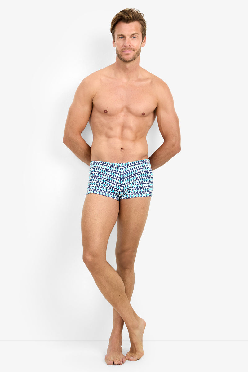 A shirtless man stands barefoot against a plain background, facing forward with a slight smile, arms relaxed behind his back. He is wearing the MONTEGO BAY SWIM TRUNK in Blue Diamond.