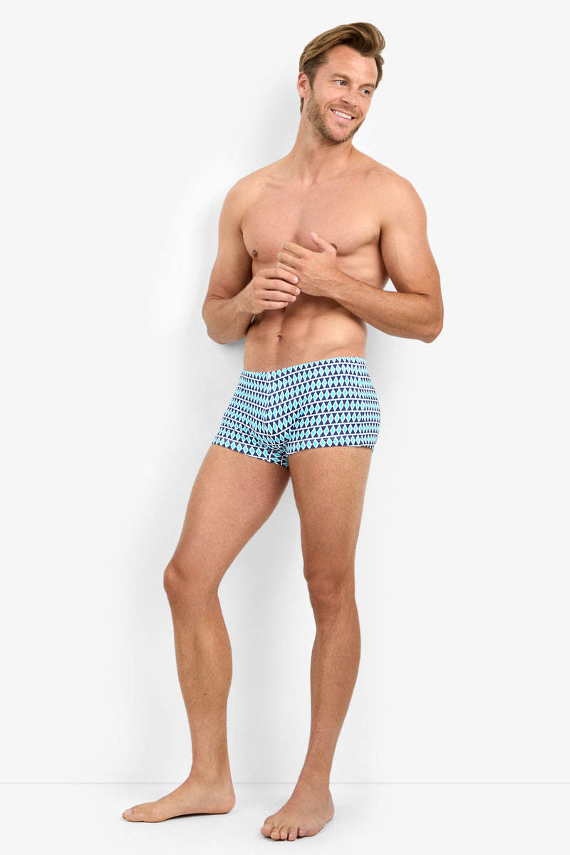 A smiling, shirtless man stands barefoot against a white background, wearing MONTEGO BAY SWIM TRUNK with blue and white patterns. He poses with one hand near his chest, looking slightly to the side.