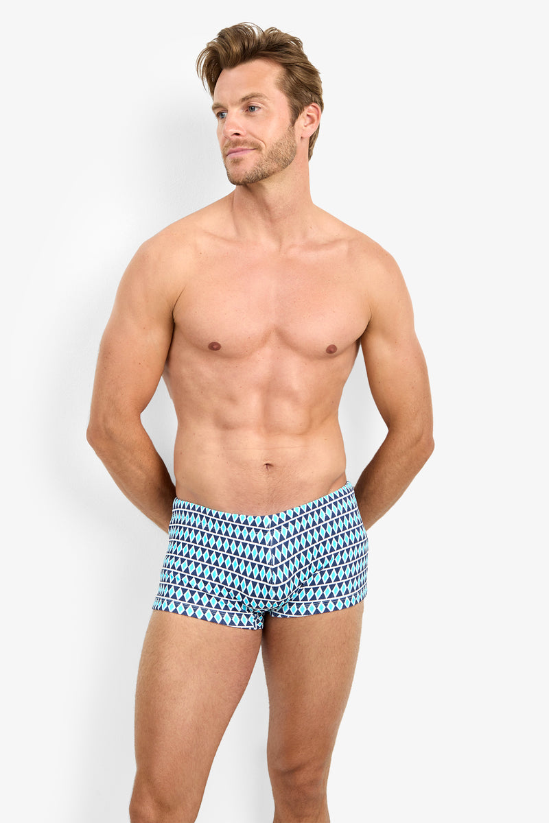 A shirtless man stands against a white background, wearing MONTEGO BAY SWIM TRUNK featuring a blue and black geometric pattern. His hands are behind his back and he gazes slightly to the side with a relaxed expression.