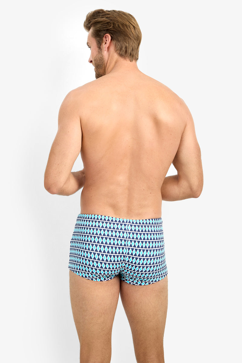 A shirtless man with light skin and short hair is seen from the back, modeling MONTEGO BAY SWIM TRUNK featuring a blue, black, and white geometric pattern, against a plain white background.